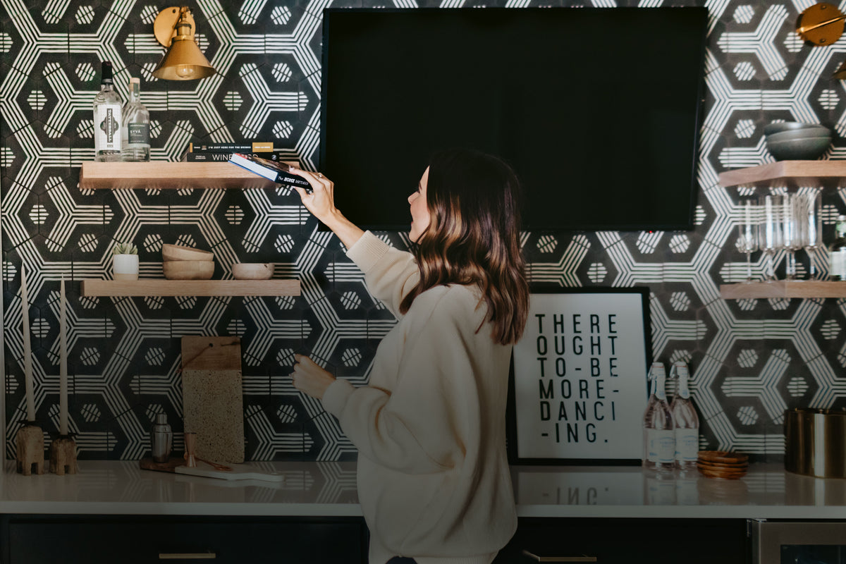 Woman arranging decor on a wall shelf beneath a television, surrounded by patterned wallpaper, dishes, bottles, and kitchenware