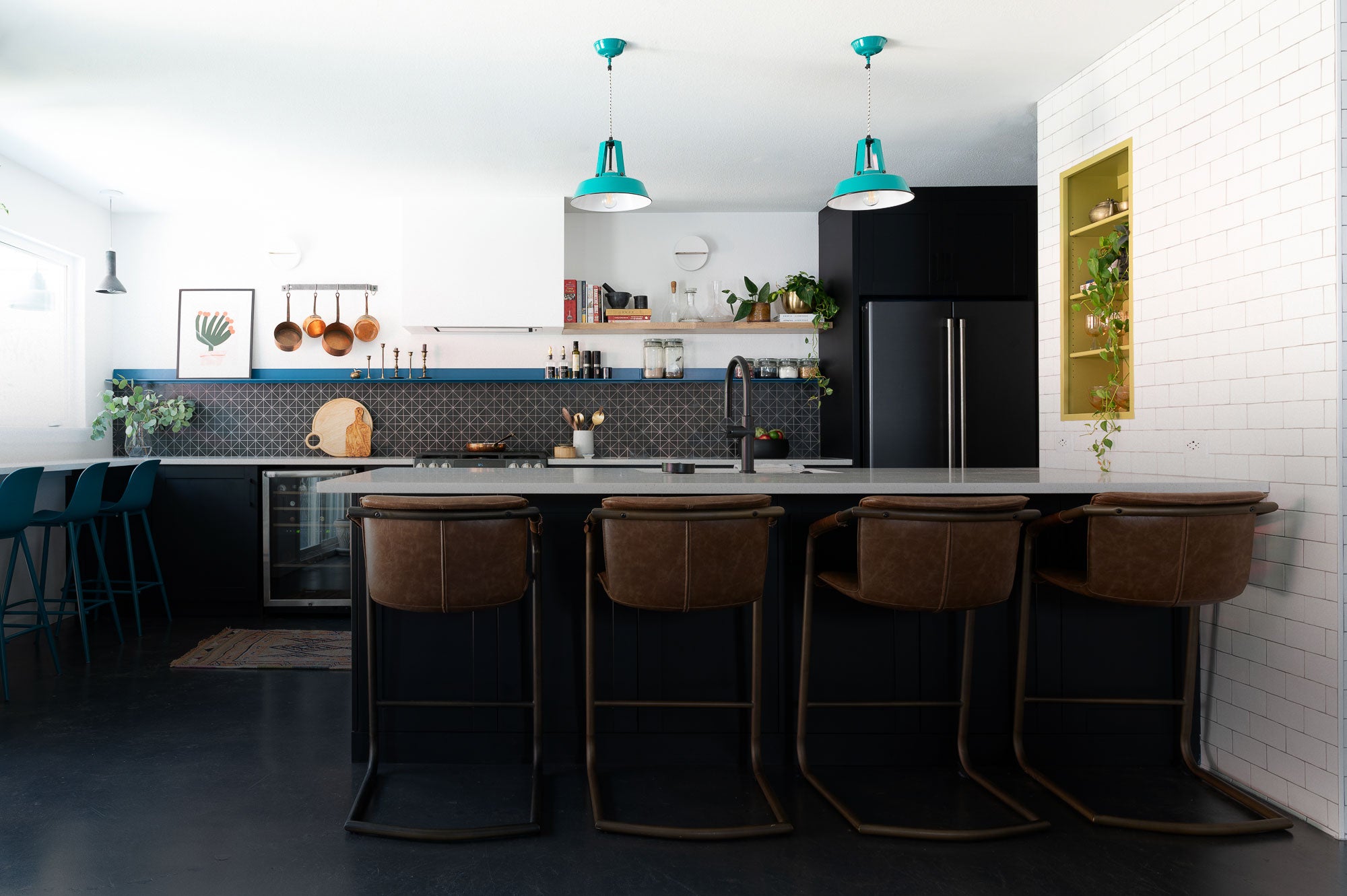 Contemporary kitchen with dark cabinetry and island topped by four brown cushioned stools, accented by teal pendant lights and open wooden shelves displaying cookbooks and plants.