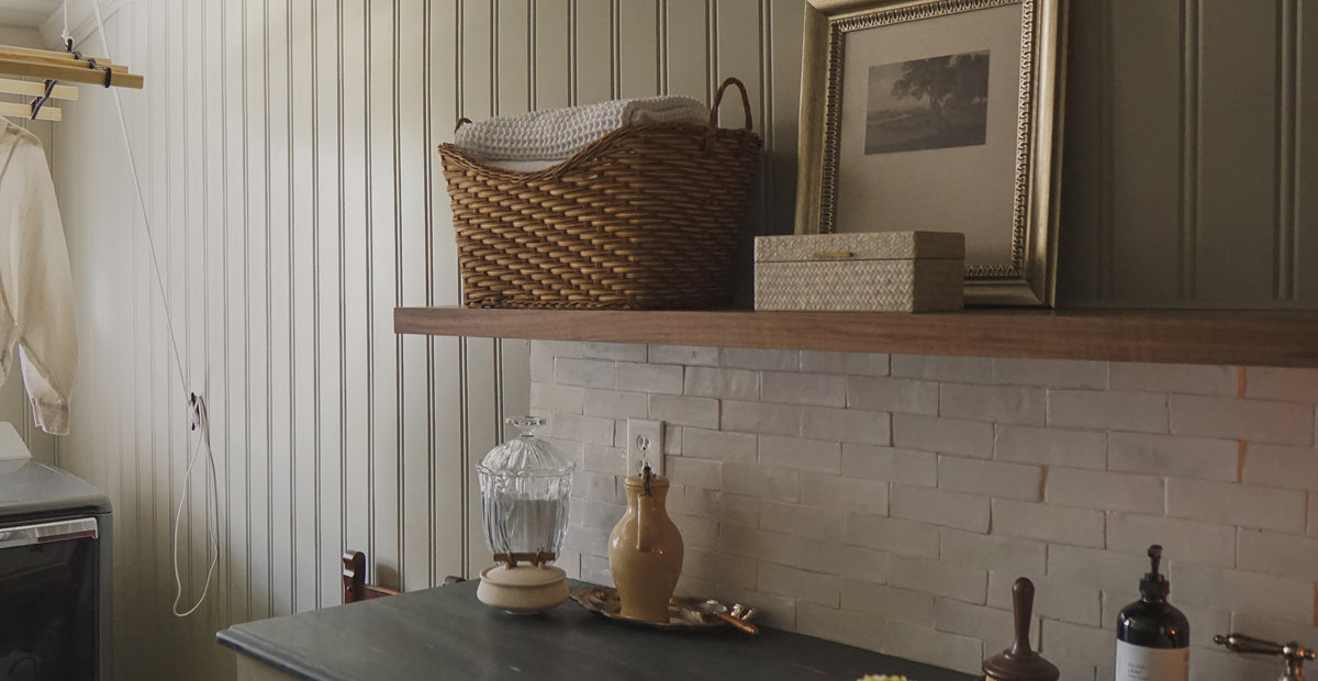 Wood floating shelf above gray brick backsplash holding framed photo, wicker basket, and storage boxes by window with curtain