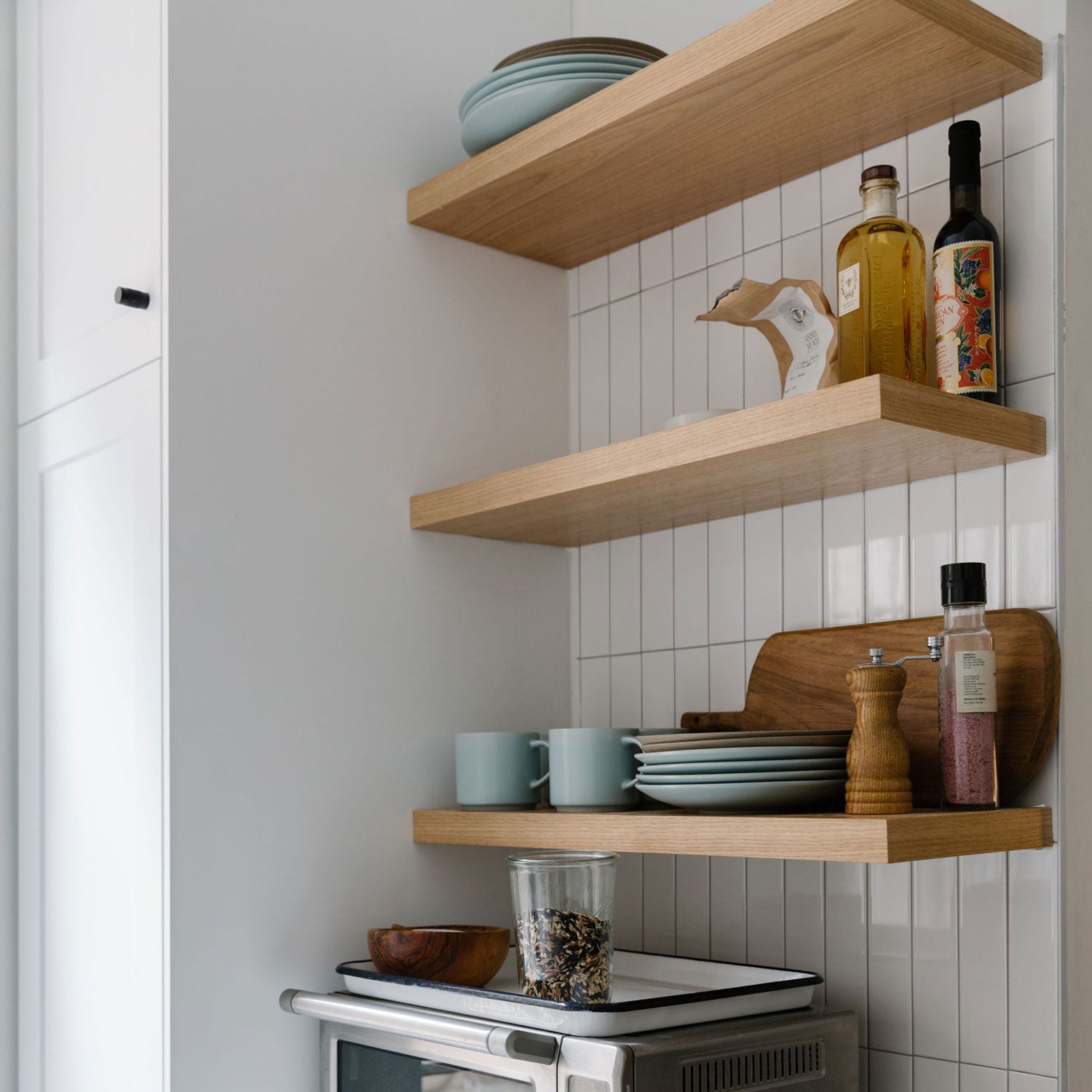Light oak floating shelves with plates, mugs, glass jar, cutting board, and bottles against white tiled kitchen wall.