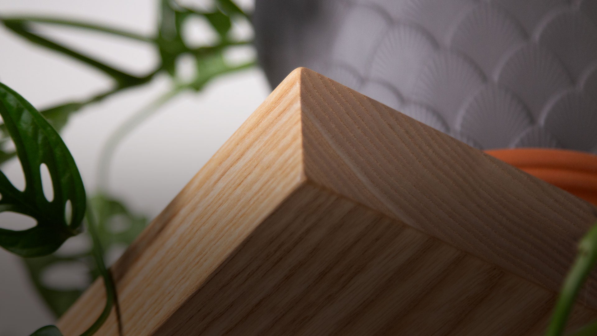 Close-up view of a light natural wood floating shelf corner surrounded by green plant leaves and a gray textured planter in soft natural light.