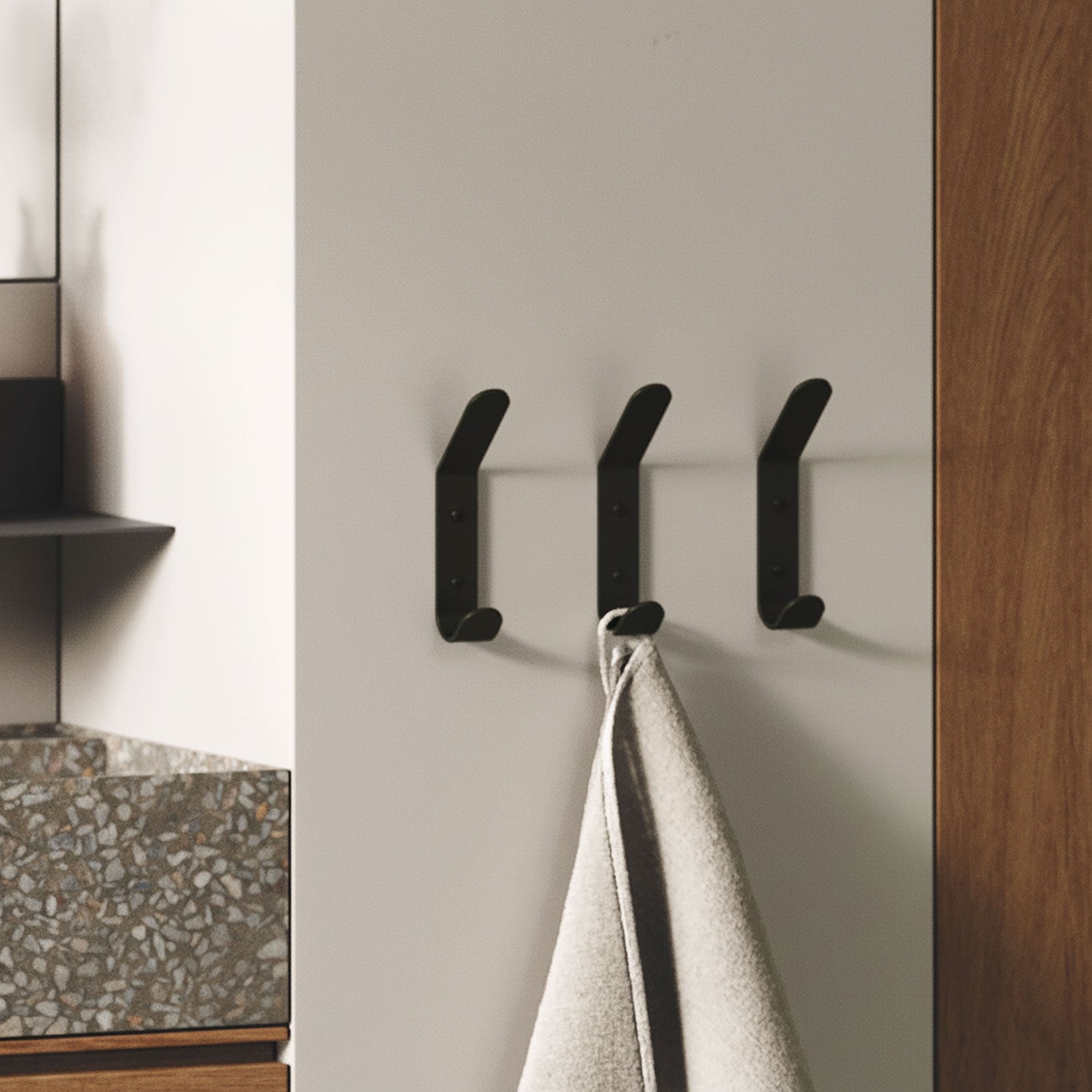 Three matte black wall hooks mounted on a light gray wall, one holding a light-colored towel, next to a terrazzo countertop and wooden cabinetry.