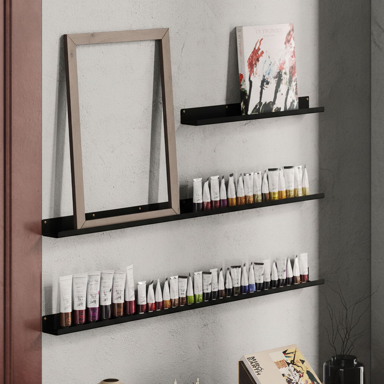 Three black metal floating wall shelves mounted on a light gray wall, holding framed art and neatly arranged tubes of paint, with a minimalistic and organized aesthetic.