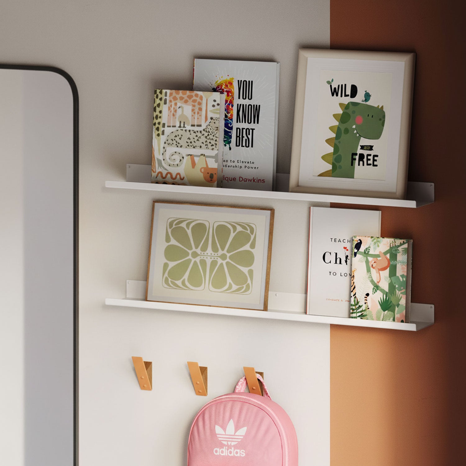 Two white floating wall shelves mounted on a beige and terracotta two-tone wall, displaying framed art prints, books, and a small notebook, with three wooden wall hooks below holding a pink Adidas backpack.