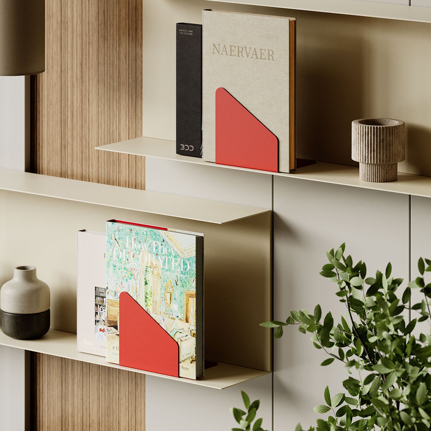 Minimalist wall shelves displaying books held upright by red metal bookends, accompanied by a small ceramic vase and a textured decor piece with a green plant nearby.