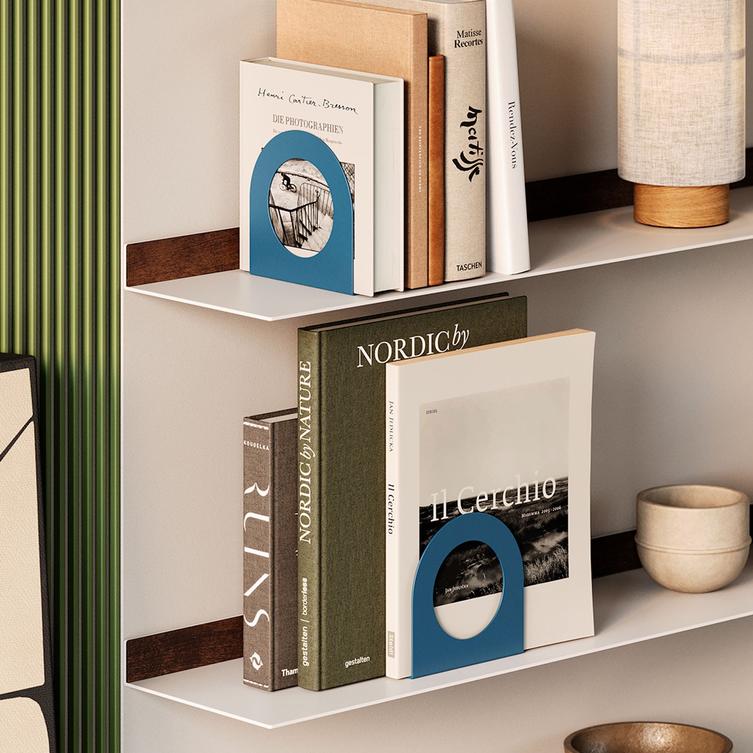 Minimalist wall shelves holding books secured with blue metal bookends, next to a fabric lamp and small pottery pieces on a neutral background.