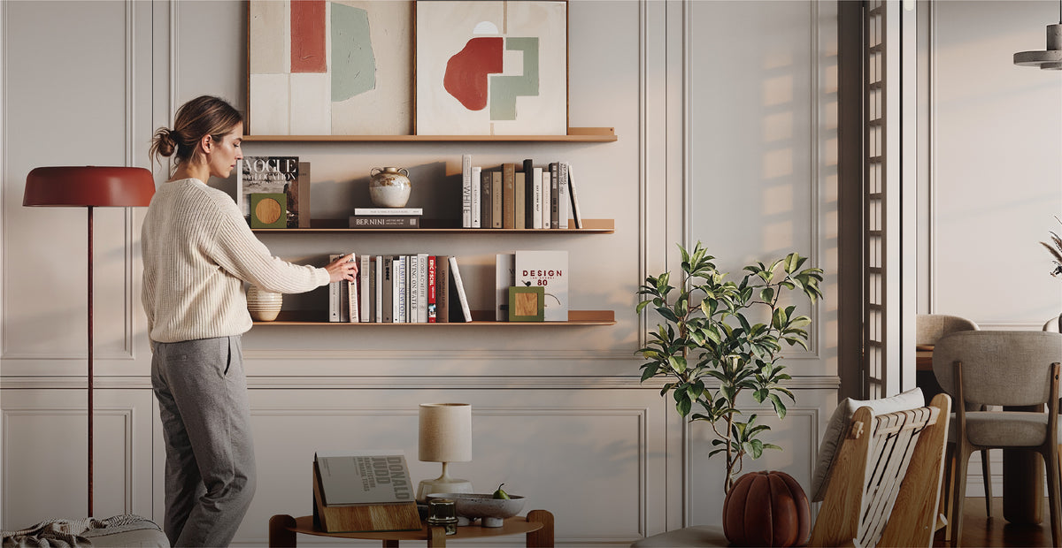 Woman arranging books on two wooden wall shelves in a bright living room with framed art plants and modern decor