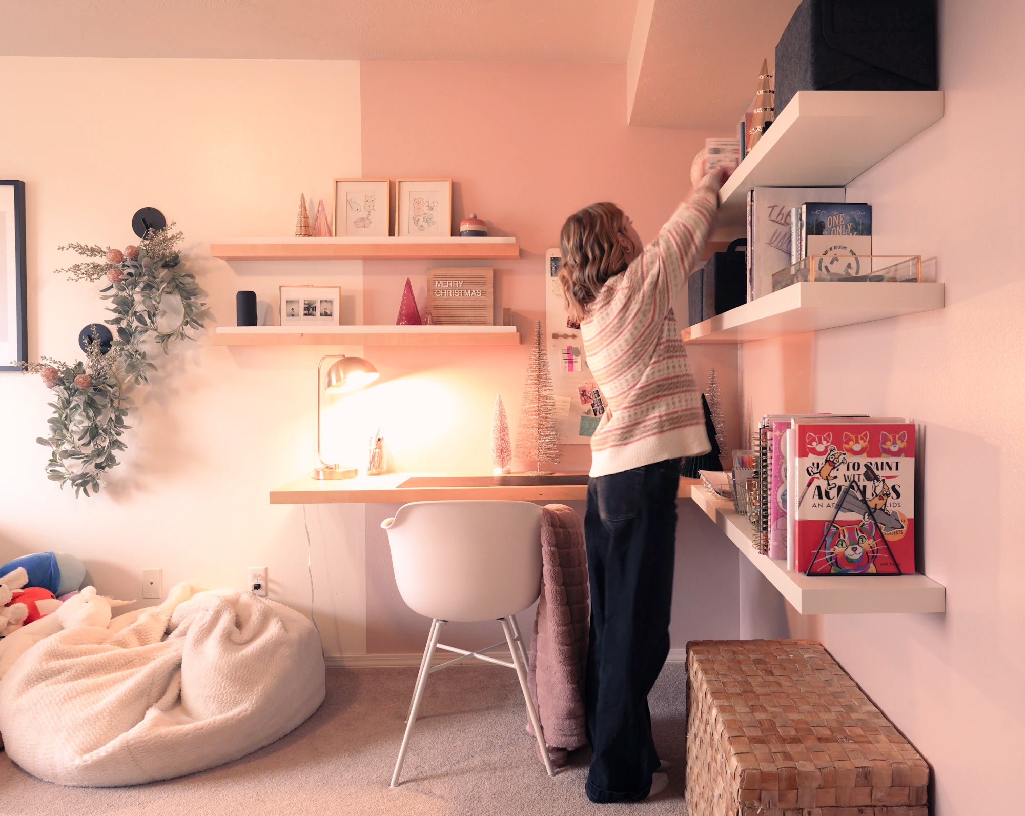 Woman reaching for books on white floating wall shelves in cozy pink-toned room with desk and bean bag chair