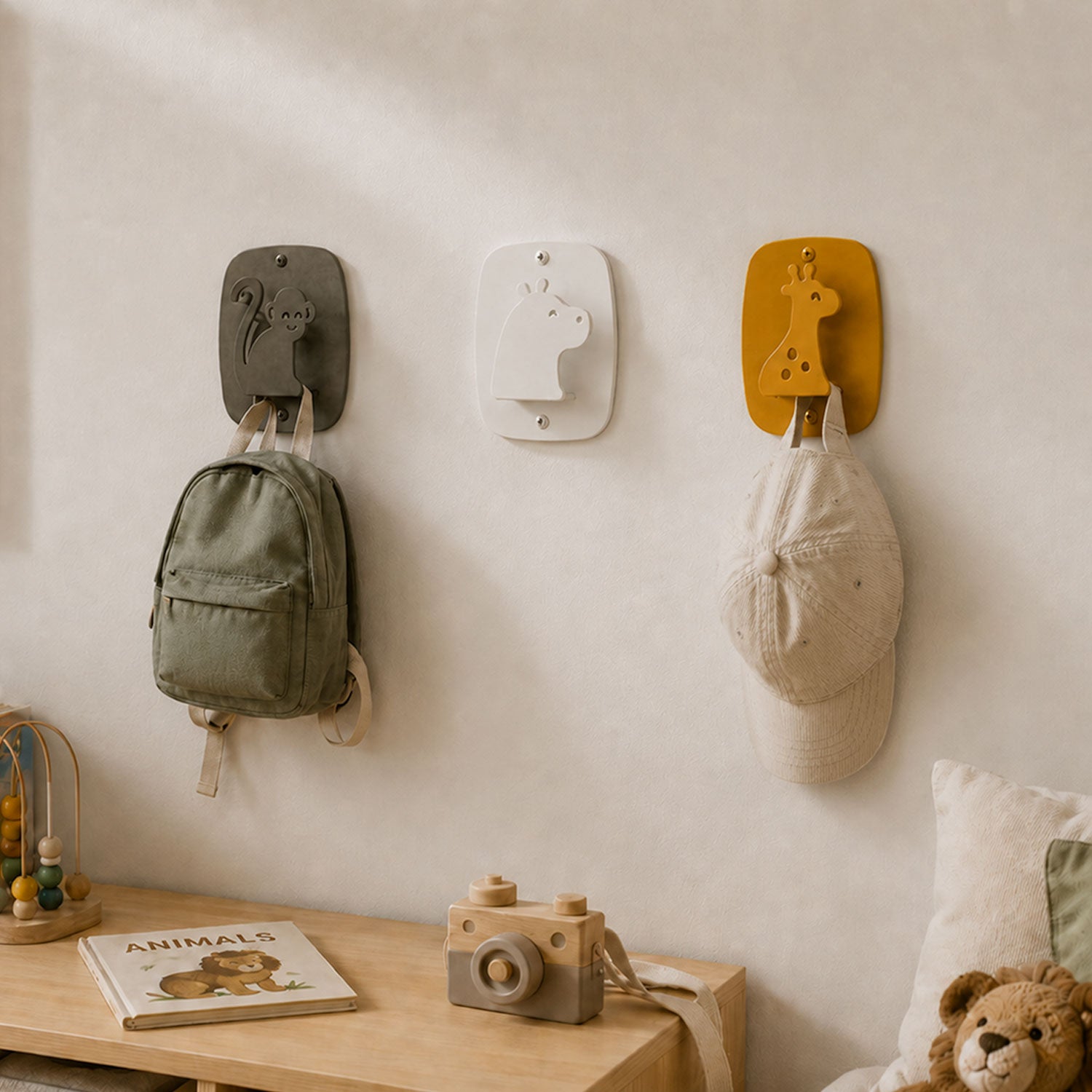 Three Kiddo Hooks mounted on a wall in a children's room, featuring playful animal designs holding a backpack and a cap above a wooden shelf with toys and a book.