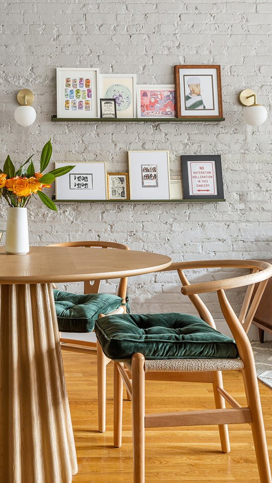 Two slim metal floating ledges display framed art against a white painted brick wall above a wood dining table.
