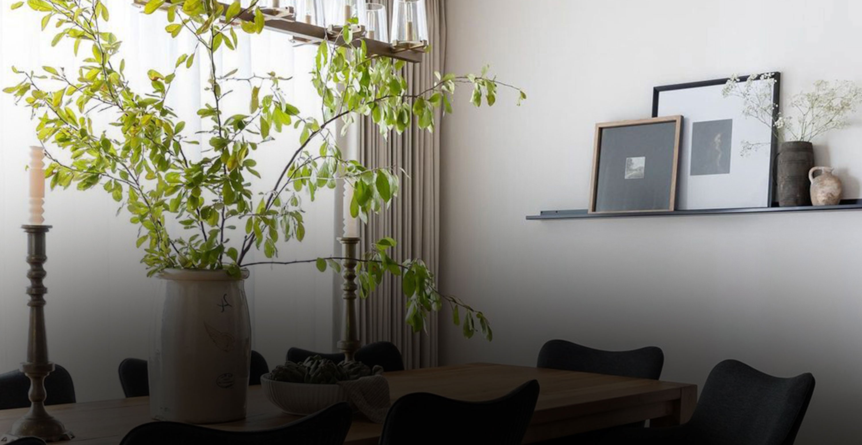 Dining room with a wooden table and black chairs, featuring a minimalist black floating shelf holding framed art and vases, and a large leafy branch centerpiece.