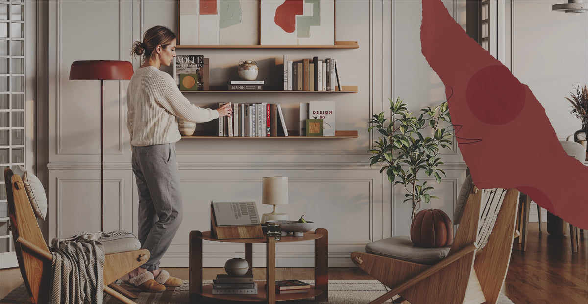 A woman arranges books on wood wall shelves in a cozy living room with framed art, plants, lounge chairs, and a coffee table