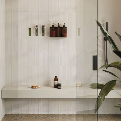 Soap Opera Shower Dispenser mounted on a white tiled shower wall with dark bottles, minimalist green hooks, and a beige floating bench below.