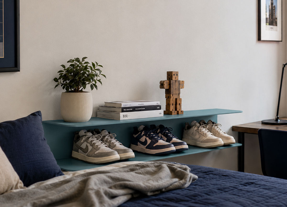 Minimalist teal floating wall shelf displaying sneakers, books, a wooden sculpture, and a potted plant in a modern bedroom setting.
