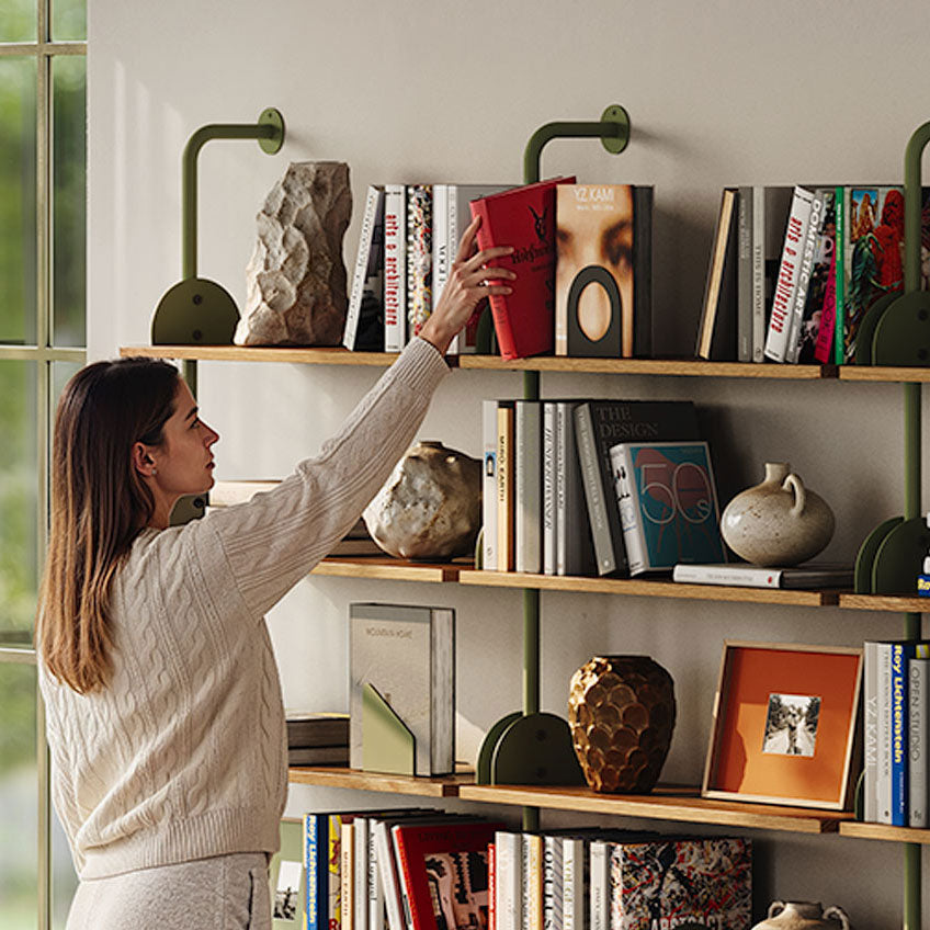 Woman arranging books and decor on wall mounted wood shelves with green metal brackets in a bright living space