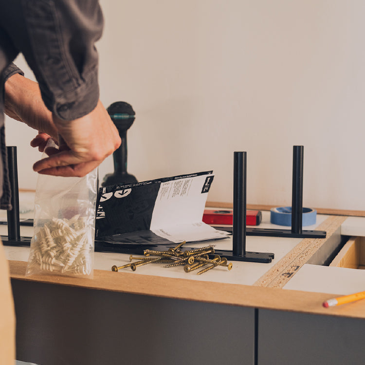 Person opening bag of white plastic wall anchors near scattered screws and metal brackets on wood surface during installation