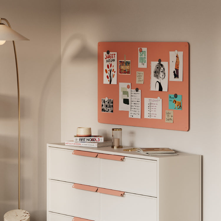 Muted coral magnetic board displaying pinned notes and artwork above a white dresser with matching accents in soft lighting