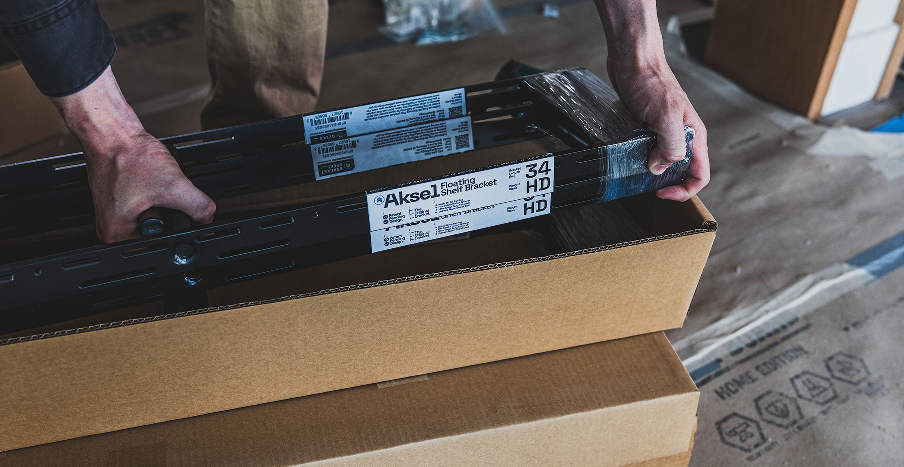 Hands unpacking black metal floating shelf hardware from a cardboard box on a wooden floor surface