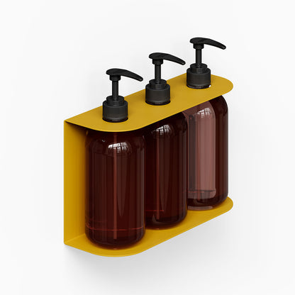 Soap Opera Shower Dispenser mounted on wall with three dark amber pump bottles in a mustard yellow minimalist metal holder designed for bathroom organization.