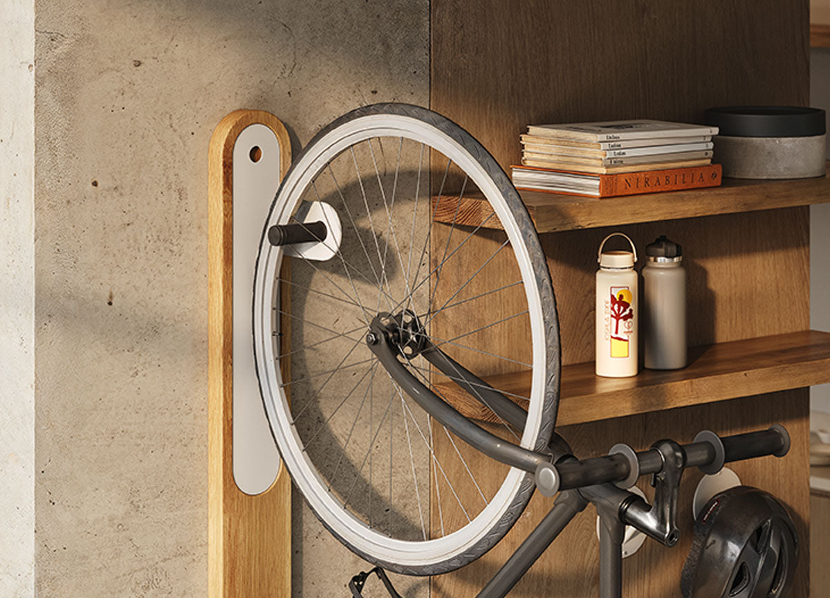 Tall wooden bike hook with white metal insert holding a bicycle front wheel near wood floating shelves with books and bottles